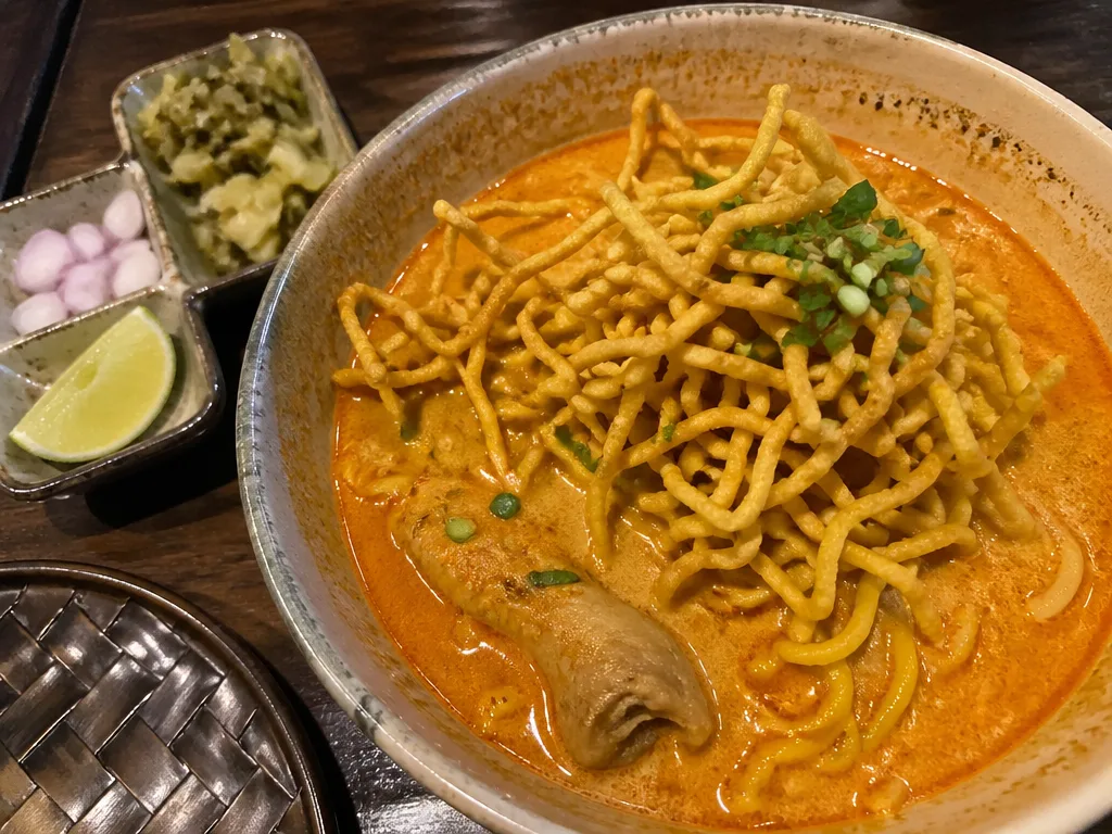 Bowl of khao soi with crispy noodles, lime, and pickled mustard greens on a wooden table