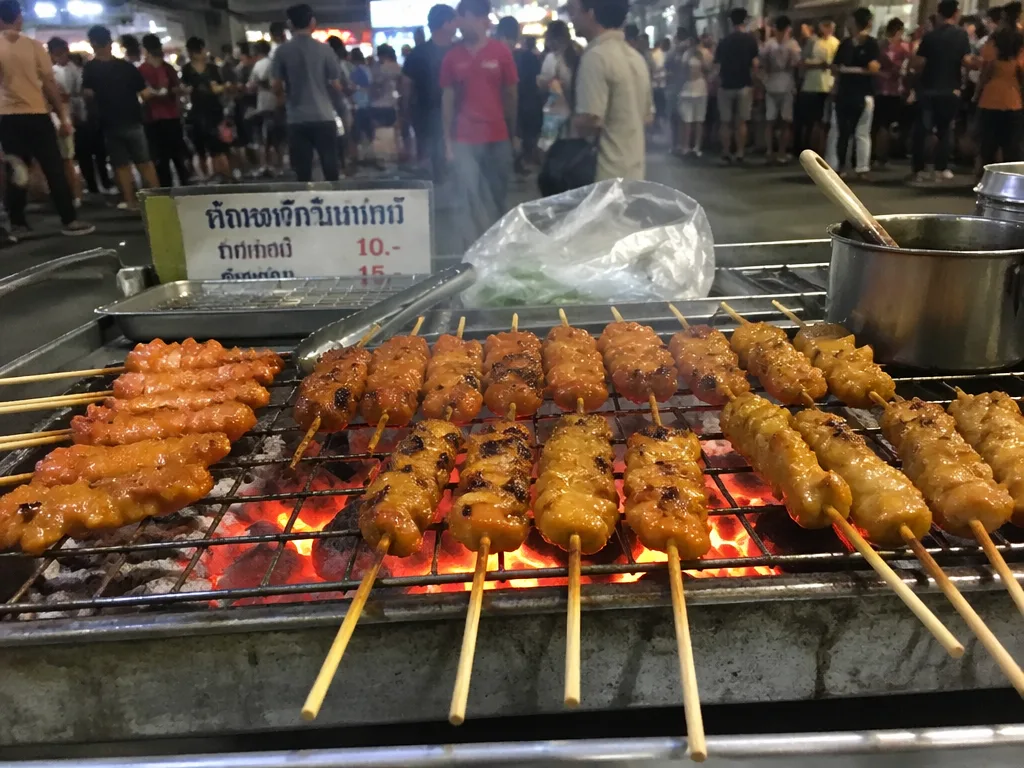 Grilled meat skewers smoking over charcoal on a Khao San Road street vendor cart