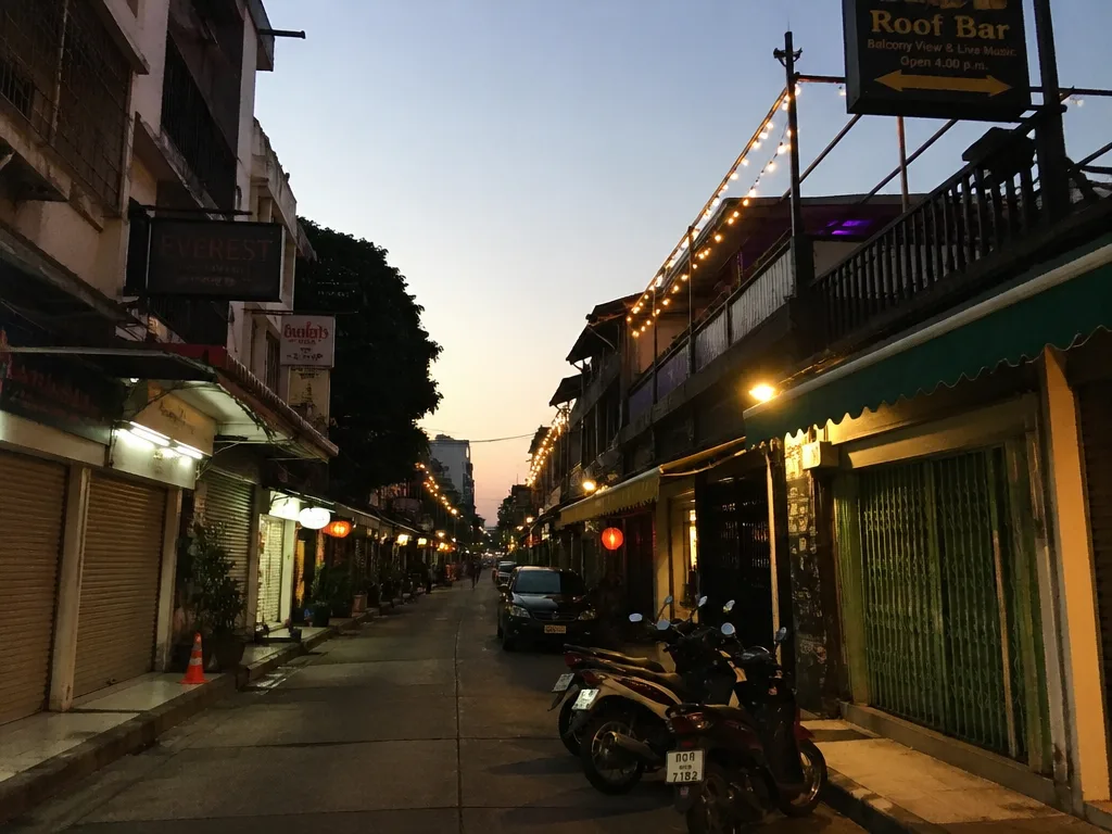Quiet Rambuttri side street with rooftop bar string lights and parked scooters in the early evening
